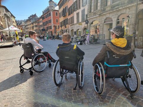Trois personnes à mobilité réduite dans des fauteuils roulants de dos sur la Place Saint Léger